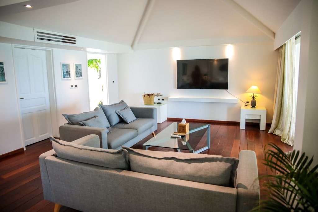 A modern living room with two gray sofas, a glass coffee table, a wall-mounted TV, wooden floors, white walls, and a side table with a lamp near a large window with light curtains.