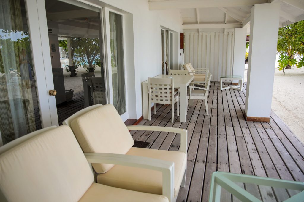 A shaded wooden patio with white cushioned chairs and tables outside a building with large glass doors and windows, overlooking a sandy area and tropical greenery.