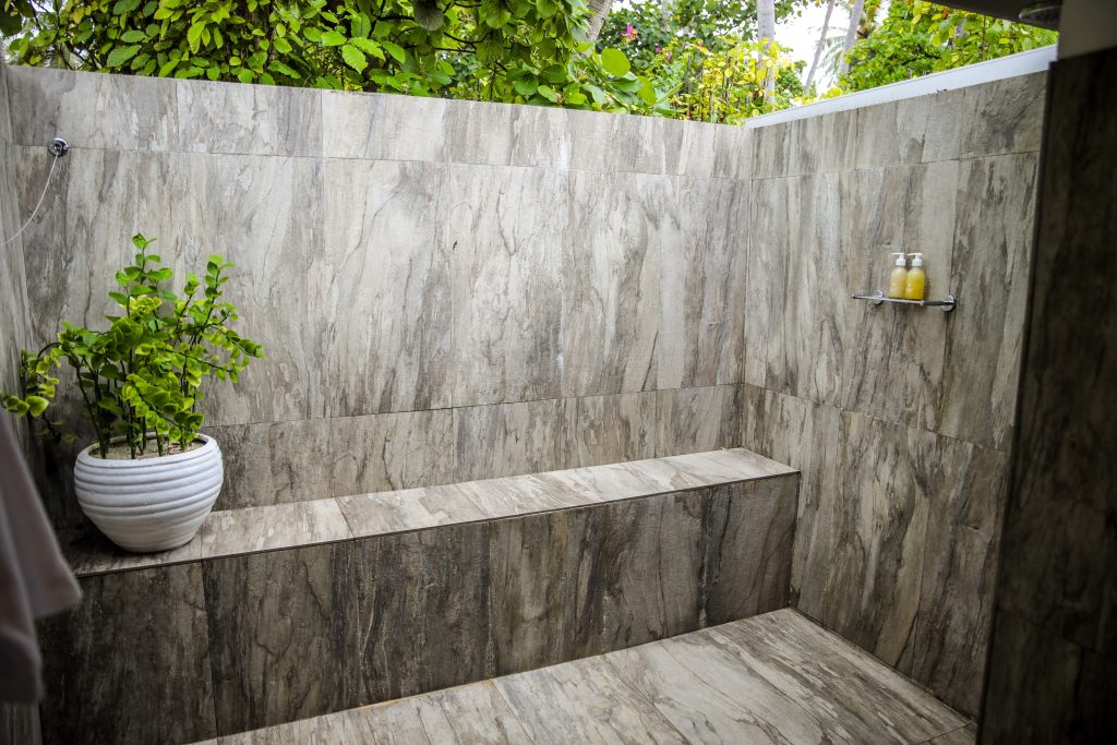 An outdoor shower with beige and gray stone walls and floors, featuring a built-in bench, a white potted plant, and two bottles on a small wall shelf. Green foliage is visible above the shower walls.
