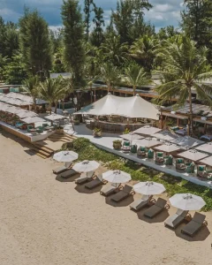 Aerial view of a tropical beach club with lounge chairs and umbrellas on sandy beach, a shaded dining area, and palm trees surrounding the open-air buildings.