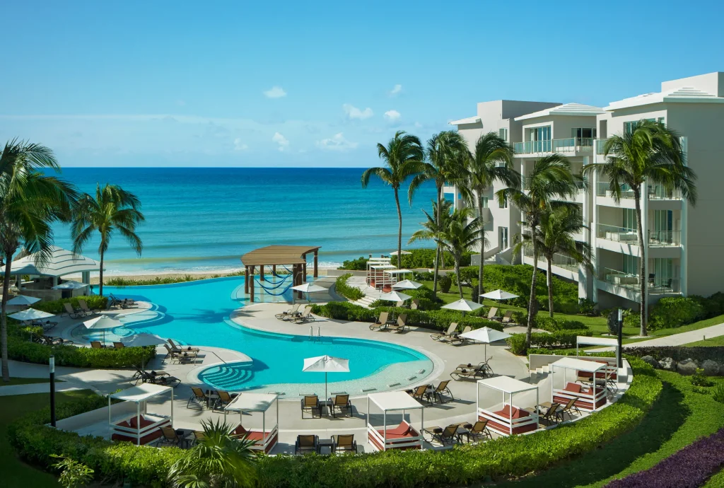 A tropical beachfront resort with palm trees, a winding pool surrounded by lounge chairs and umbrellas, and modern white buildings overlooking a calm, turquoise ocean under a clear blue sky.