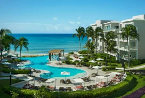 A tropical beachfront resort with palm trees, a winding pool surrounded by lounge chairs and umbrellas, and modern white buildings overlooking a calm, turquoise ocean under a clear blue sky.