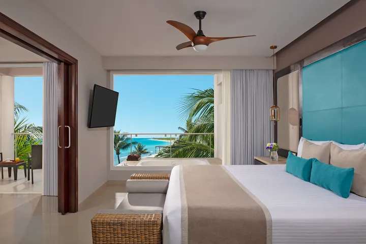 Modern hotel room with a large bed, blue and beige decor, ceiling fan, and a balcony offering a view of palm trees and the ocean under a clear blue sky. A TV is mounted near the bed, and patio furniture is visible outside.
