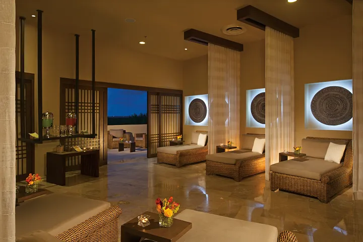 A modern spa lounge with wicker chaise lounges, sheer curtains, soft lighting, and decorative wall art. Small tables hold flowers and candles, and there is a refreshment station on the left. An outdoor area is visible in the background.