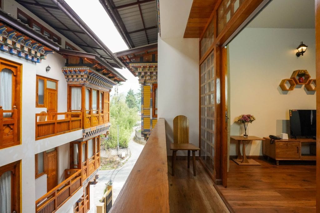 A balcony view of a traditional-style building with wooden accents and decorative patterns, facing another similar building; a cozy room with a chair, TV, and flowers is visible through a sliding door.