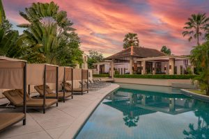 A luxurious poolside area with lounge chairs and canopy beds beside a calm swimming pool, surrounded by tropical plants, with a modern villa and vibrant pink-orange sunset sky in the background.
