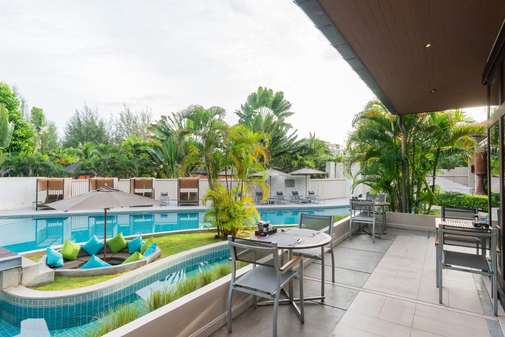 Outdoor restaurant patio overlooking a modern swimming pool with colorful cushions, potted plants, and tropical greenery. Tables and chairs are set for dining, creating a relaxing, resort-like atmosphere.