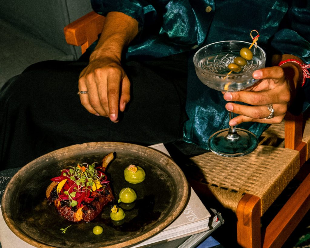 A person in a dark outfit sits on a wooden chair, holding a cocktail glass with olives. A plate with a gourmet dish and green garnish rests on their lap, along with several green fruits on the side.