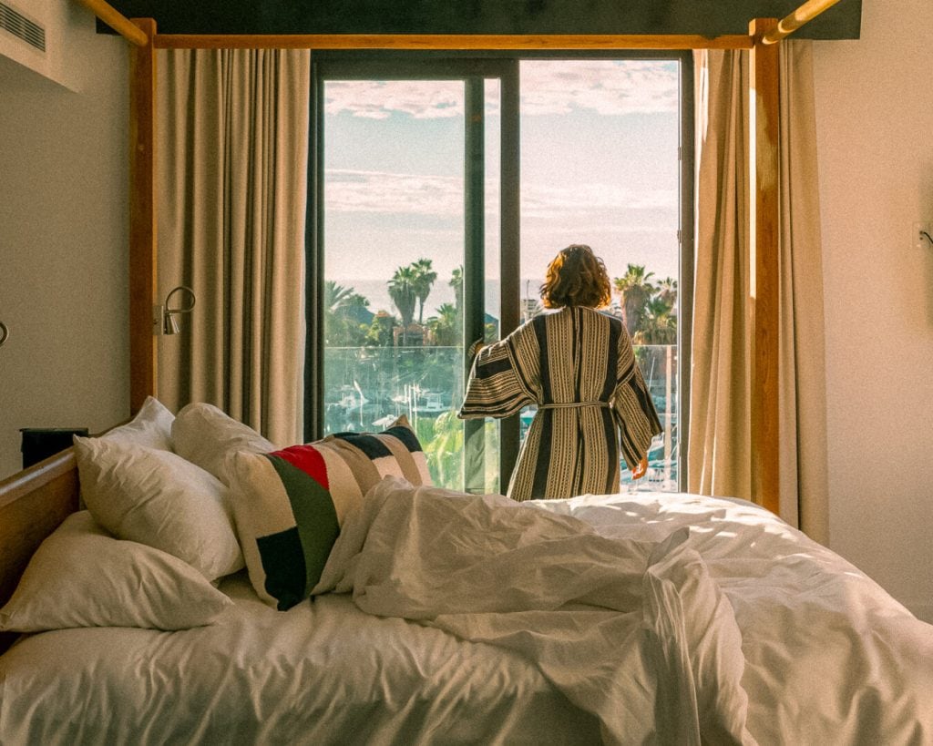 A person in a robe stands on a balcony, looking out at palm trees and a sunny sky. The rumpled bed with pillows and sunlight streaming in suggests a relaxing morning in a cozy hotel room.
