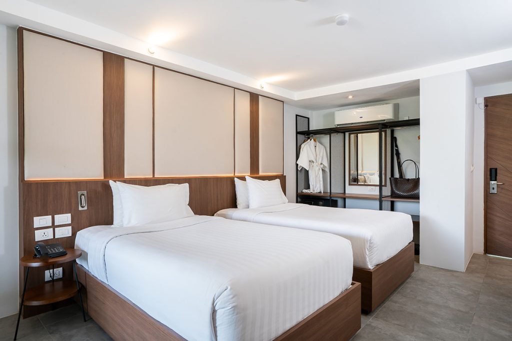 A modern hotel room with two neatly made single beds, white bedding, wood accents, a wall-mounted wardrobe with hangers and robes, and a door in the background. The room is bright and tidy.