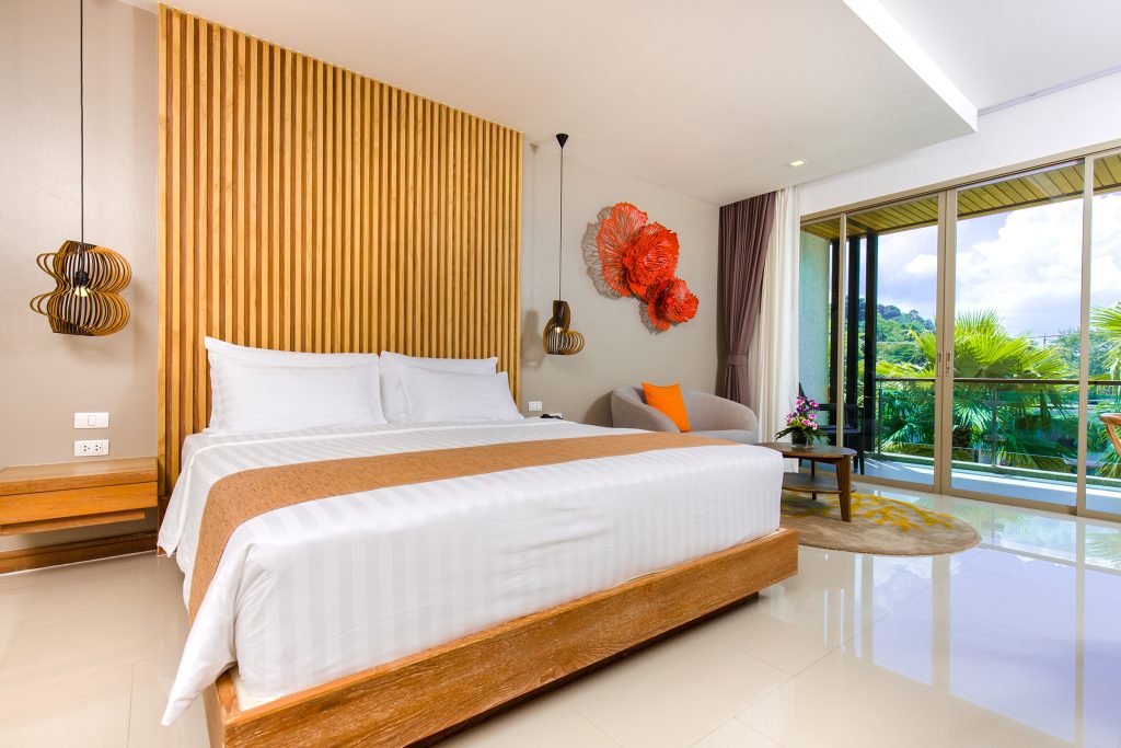 A modern hotel room with a large bed, white linens, wood accents, unique pendant lights, orange decor on the wall, a gray chair, and a balcony overlooking greenery and trees outside.