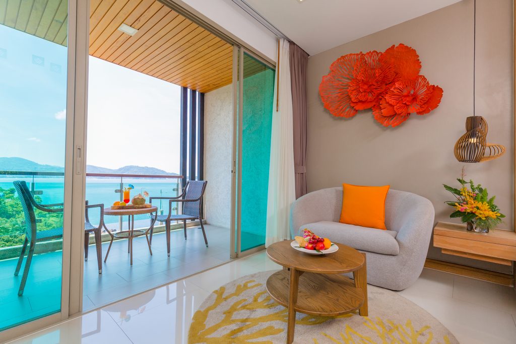 A modern living room with a gray armchair, orange pillow, wooden table with fruit, and wall art. Sliding glass doors open to a balcony with a table, chairs, and a scenic view of water and hills.