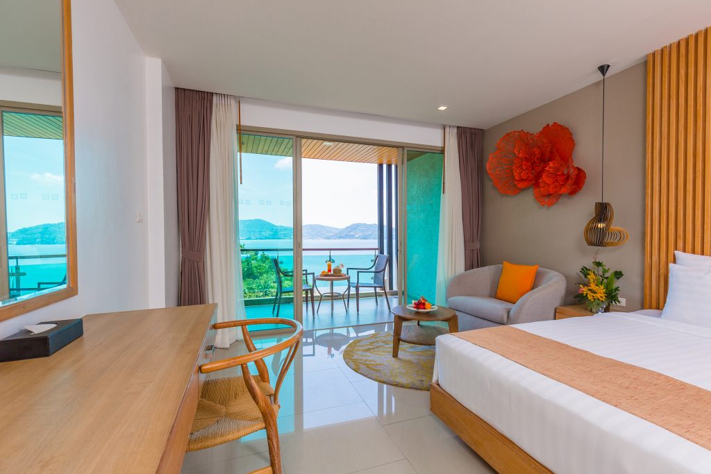 A modern hotel room with a bed, sofa, desk, and chair, decorated in neutral tones with orange accents. Glass doors open to a balcony with seating and a view of the ocean and distant hills.