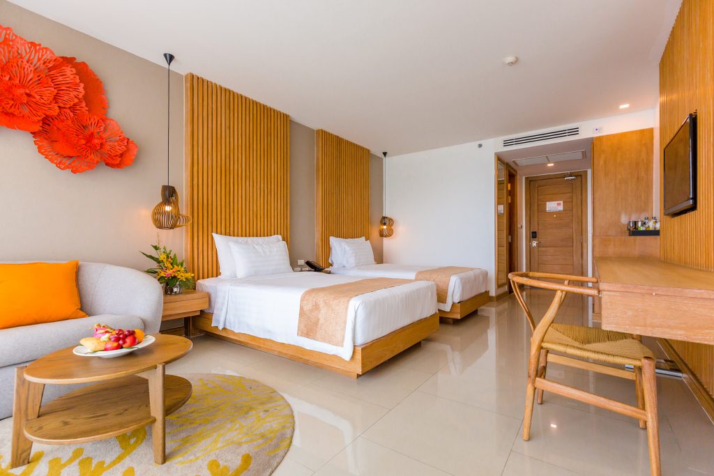A modern hotel room with two twin beds, wooden accents, a desk with a chair, a sofa with an orange pillow, a round coffee table with fruit, and bright wall art. The room is clean and well-lit with natural light.
