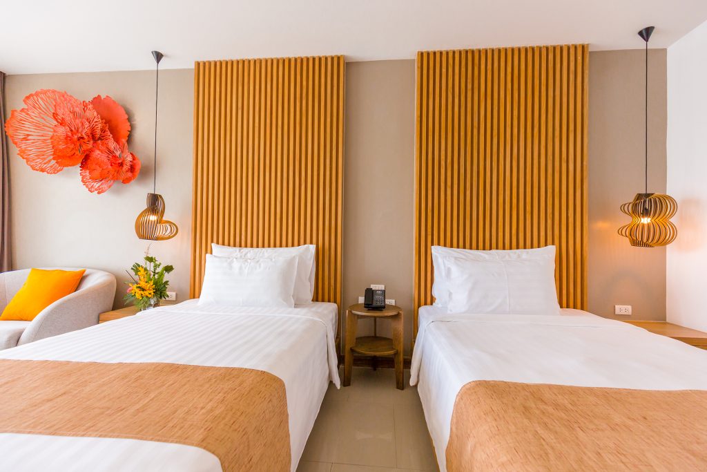 Two neatly made single beds with white linens and light brown throws, separated by a small nightstand. Warm lighting, orange accent panels, modern pendant lamps, and red wall art decorate the contemporary room.