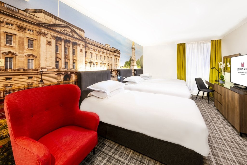 Hotel room with three single beds, a red armchair, desk with chair, TV, and a large mural of a historic building on the wall. The room is bright with white bedding and green curtains by the window.