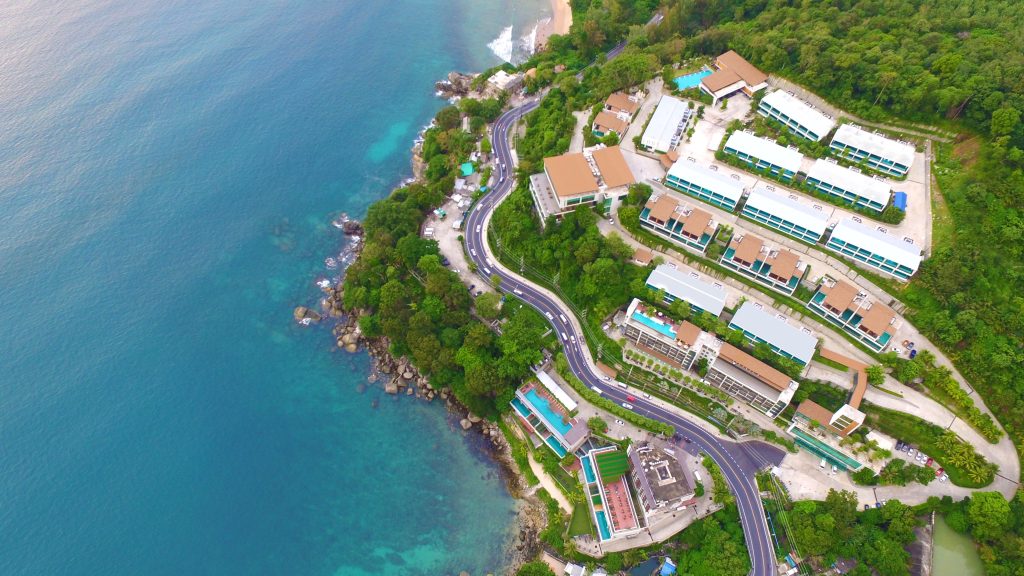 Aerial view of a coastal area with winding roads, modern buildings, and lush greenery beside clear blue ocean water and a rocky shoreline.
