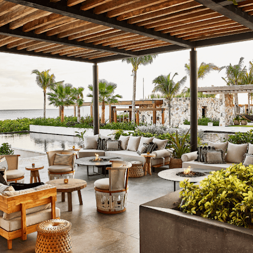 Open-air lounge with cushioned chairs and tables under a wooden pergola, overlooking a pool, palm trees, and modern stone buildings with ocean visible in the background.