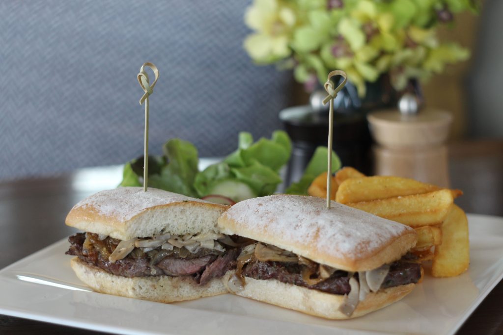 A steak sandwich with caramelized onions is cut in half and served on a square white plate with thick-cut fries and a small side salad. Skewers hold the sandwich halves together. A blurred background shows condiments and flowers.