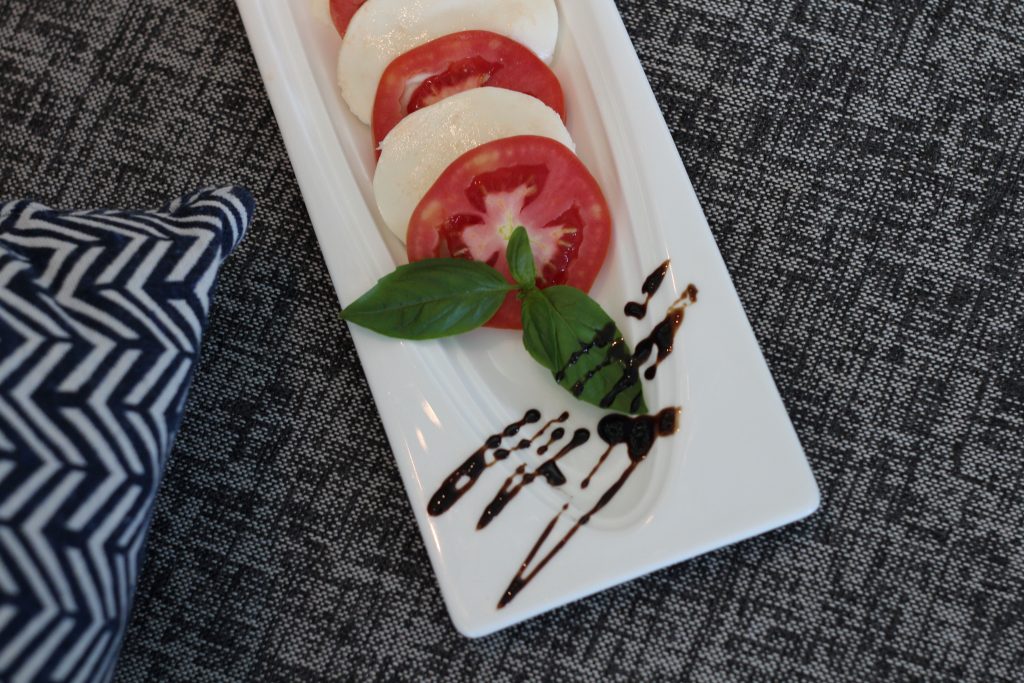 A rectangular white plate with sliced tomatoes and mozzarella, garnished with fresh basil leaves and a drizzle of balsamic glaze, on a textured gray surface next to a patterned napkin.