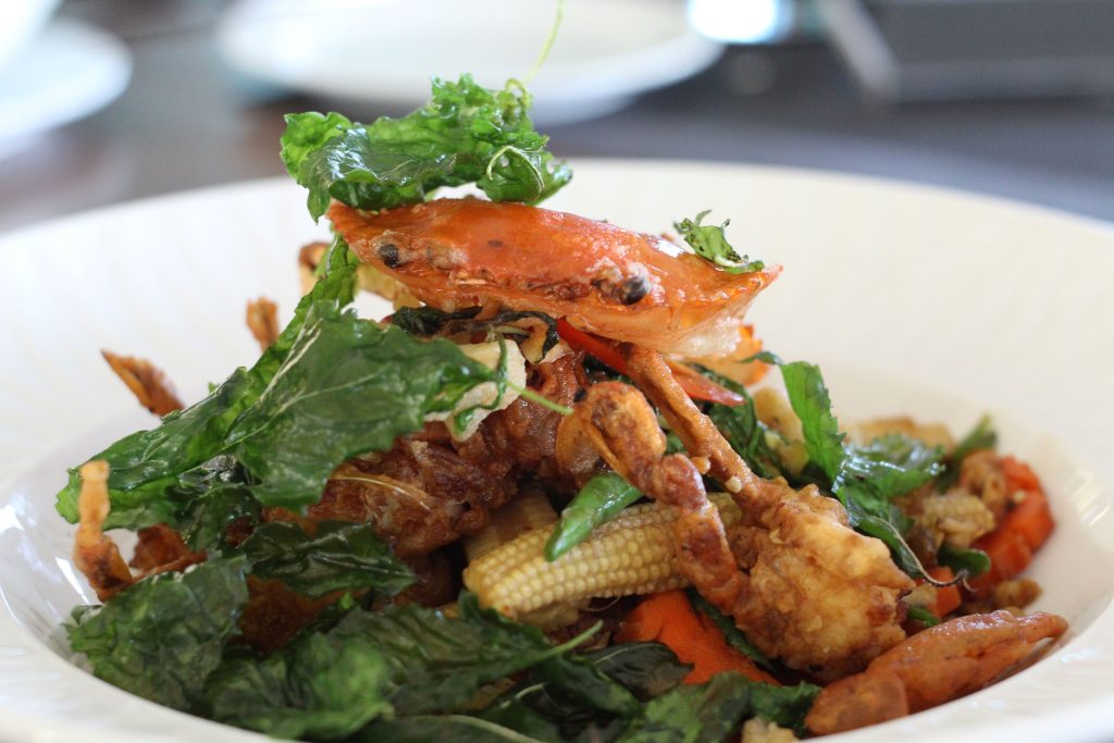 A close-up of a seafood dish featuring a cooked crab, crispy fried greens, baby corn, and vegetables, all served on a white plate.