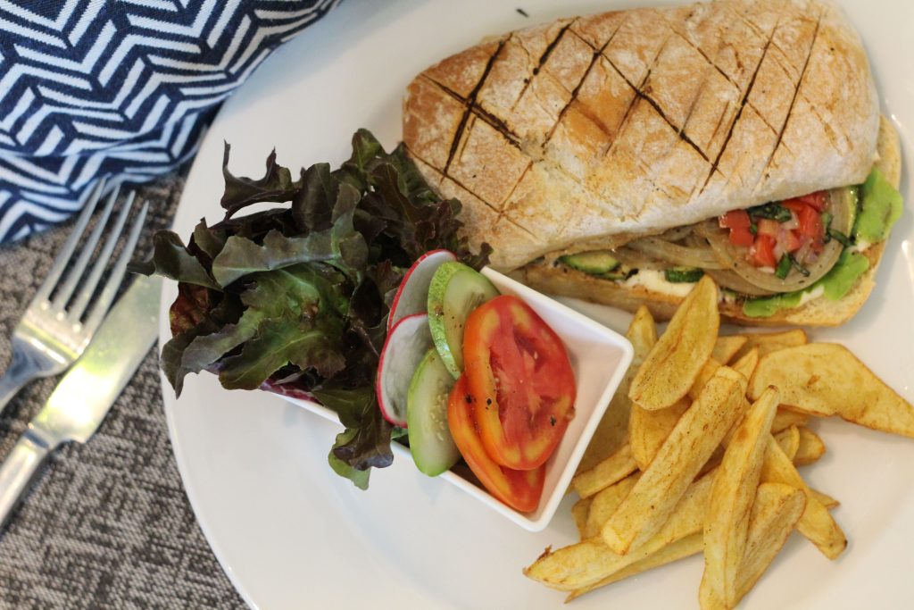 A plate with a ciabatta sandwich filled with vegetables, a side of thick-cut fries, leafy greens, and sliced cucumber and tomato. A fork and napkin are placed beside the plate.