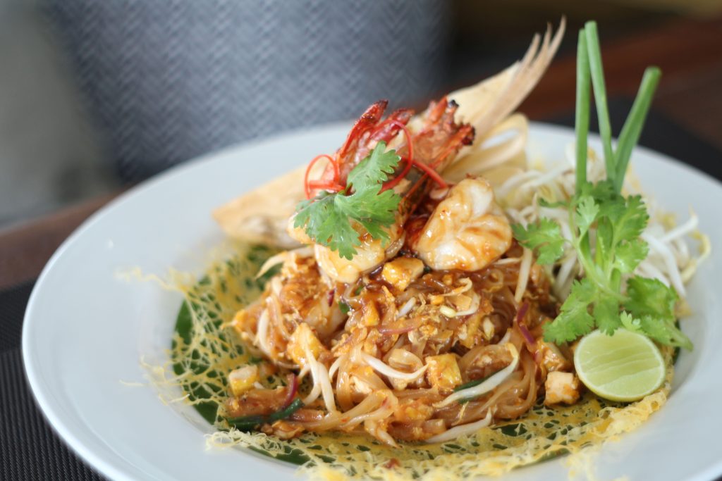 A plate of Pad Thai topped with shrimp, garnished with cilantro, chili, a lime wedge, and green onions. The dish is arranged with noodles, vegetables, and an intricate egg net on a white plate.