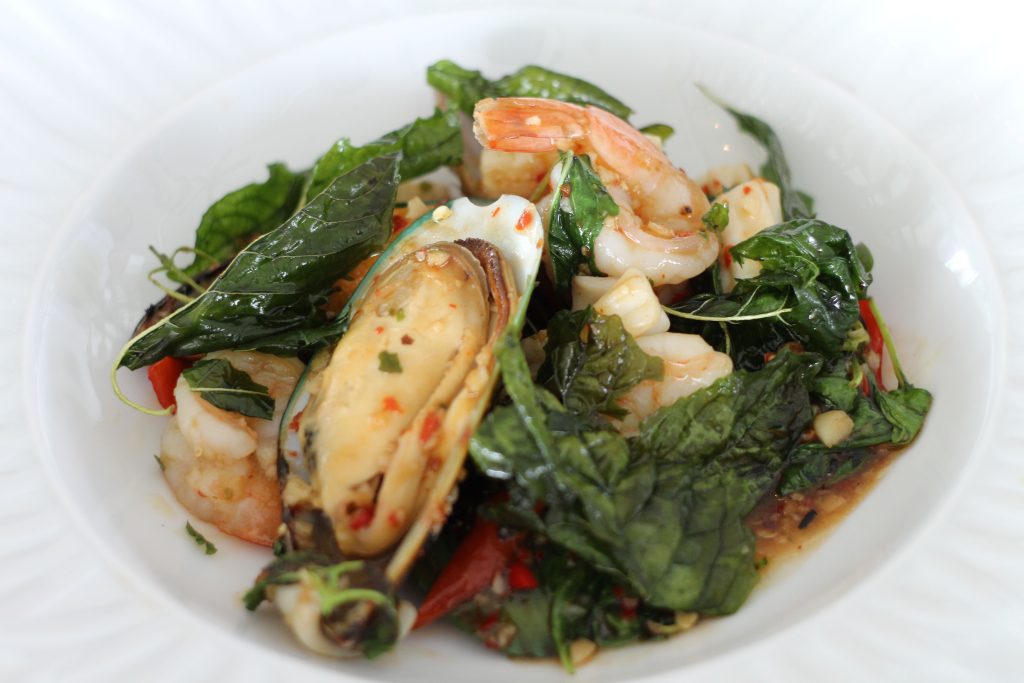 A white plate with a seafood dish featuring mussels, shrimp, leafy greens, and herbs in a light sauce. The dish appears seasoned, colorful, and garnished with fresh green leaves.