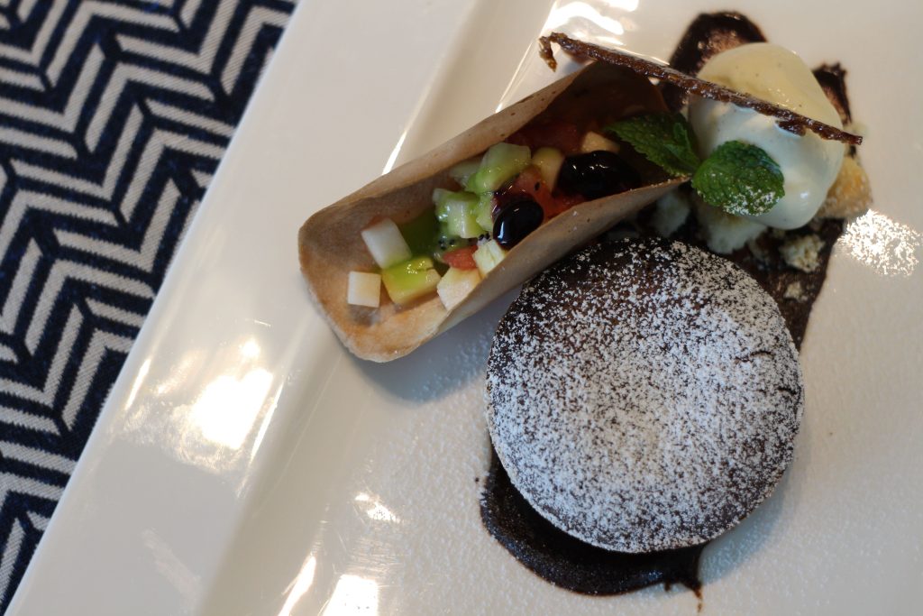 A round chocolate dessert dusted with powdered sugar sits beside a wafer filled with diced fruit, garnished with a mint leaf and a scoop of vanilla ice cream on a white rectangular plate.