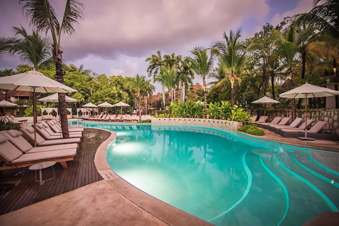 A tropical resort pool surrounded by palm trees, lounge chairs, and white umbrellas, with cloudy skies overhead and lush greenery in the background.