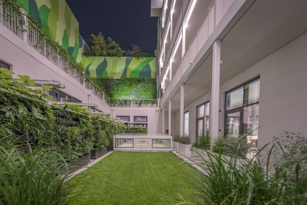A modern courtyard at night with artificial grass, lush green vertical gardens on the walls, and a mural with green shapes. The building features large windows and bright exterior lighting.