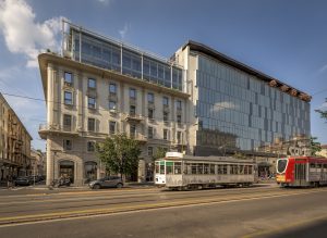 A street scene in a city with a modern glass building next to an older, ornate stone building; a white tram and a red bus travel on the road under a blue sky with scattered clouds.