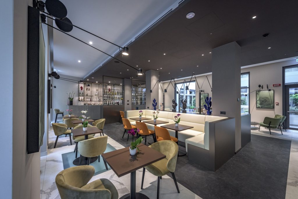 Modern restaurant interior with elegant tables, green and orange chairs, beige booth seating, decorative plants, and a sleek bar area with shelves of bottles in the background. Large windows provide natural light.