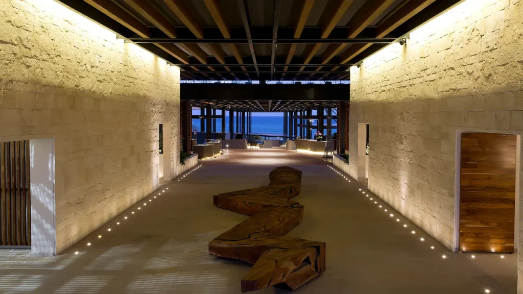A modern, open-air hallway with stone walls, wooden ceiling beams, floor lights, and a zigzag wooden sculpture leading toward a seating area with a view of the ocean in the distance.
