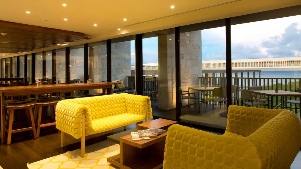 Modern lounge area with two yellow couches, wooden tables, and bar stools by large floor-to-ceiling windows overlooking an outdoor patio and water view at sunset. Magazines and décor items are on the central table.