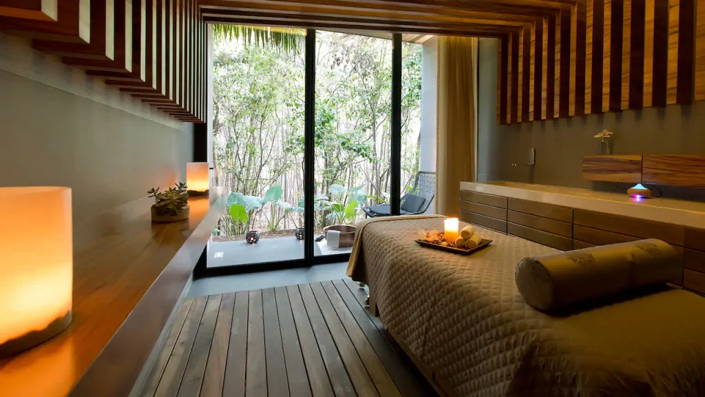 A serene spa room with a massage table, candles, wooden accents, and large glass doors opening to a lush garden outside, creating a peaceful, natural atmosphere.
