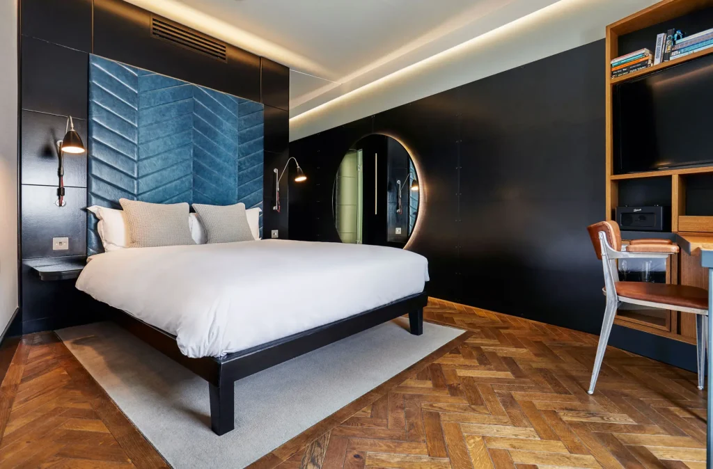 A modern hotel room with a large bed, blue padded headboard, wall-mounted reading lights, a round mirror, wooden herringbone floor, and a desk with a chair and shelving. The decor is sleek and contemporary.
