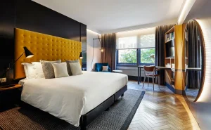 A modern hotel room with a large bed featuring a mustard yellow tufted headboard, white bedding, and decorative pillows. The room has wooden floors, a desk, a chair, and large windows letting in natural light.