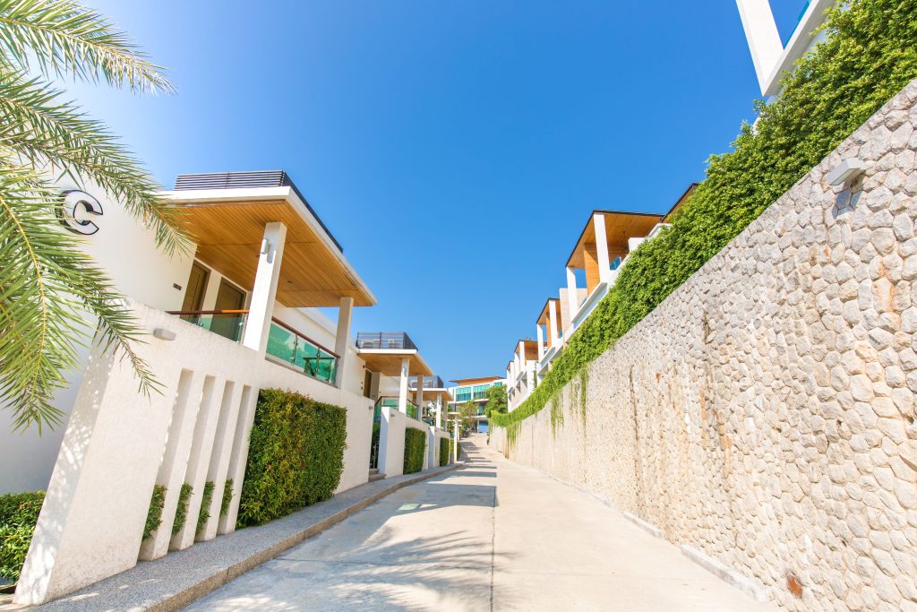 A sunny walkway lined with modern, white buildings and tall stone walls covered in green vines, beneath a clear blue sky with a palm tree on the left side.