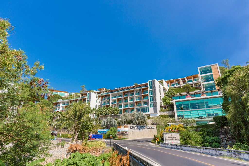 Modern multi-story resort with glass balconies and greenery on terraces, set on a hill above a road. Blue sky overhead and lush vegetation around the building create a tropical feel. Signs are visible near the entrance.