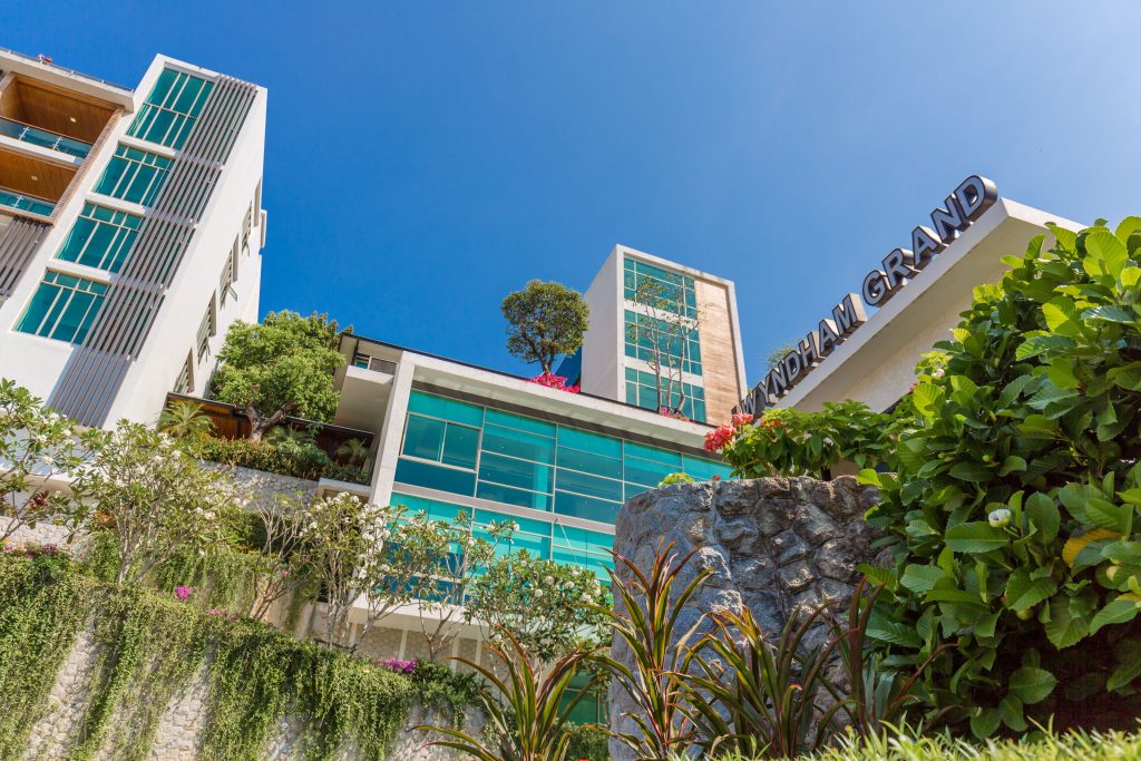 Modern hotel building with large glass windows, lush greenery, and plants in the foreground. The Wyndham Grand sign is visible on the upper right. Bright blue sky is in the background.