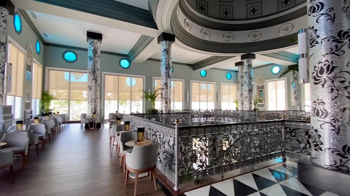 A spacious, elegantly decorated restaurant with ornate patterned columns, decorative metal railings, white and gray chairs, and large windows letting in natural light. The floor features a black and white checkered pattern.