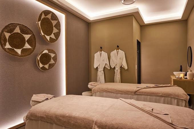A serene spa room with two massage tables, soft beige decor, woven wall baskets, two white robes hanging on hooks, dim lighting, and a small counter with spa accessories.