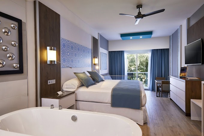 Modern hotel room with a large bed, blue and white decor, a freestanding bathtub, flat-screen TV, desk, and a window with blue curtains overlooking greenery. Ceiling fan and contemporary lighting add to the ambiance.