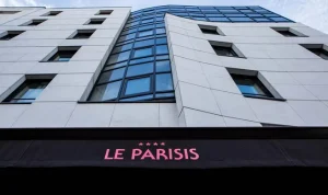 Low-angle view of the modern facade of Hotel Le Parisis, featuring geometric white panels, blue-tinted windows, and a sign with pink text reading “Le Parisis” above the entrance.