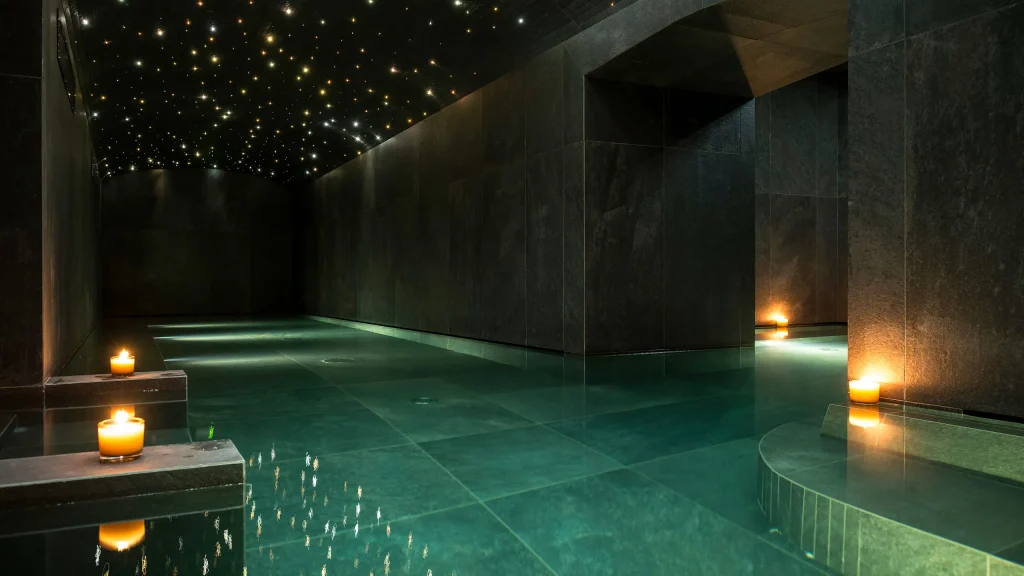A tranquil indoor spa pool with dark stone walls, floating candles on the water, and a ceiling illuminated with small, star-like lights, creating a serene and relaxing atmosphere.