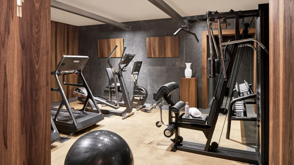 Modern home gym with treadmills, a stationary bike, a multi-use weight machine, a black exercise ball, towels, water bottles, and stylish wood and stone decor on the walls and floor.