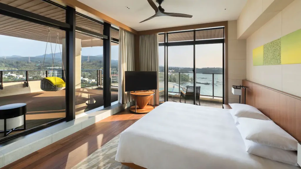 Modern hotel room with large windows, a king-sized bed, a ceiling fan, and a balcony featuring a hanging chair and outdoor seating, offering scenic views of mountains and a river.