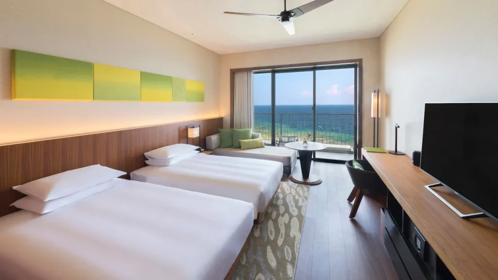 Modern hotel room with two beds, a large TV, desk, and a seating area by a window. Balcony doors open to an ocean view. The decor is minimal with green accents, wood paneling, and a ceiling fan.