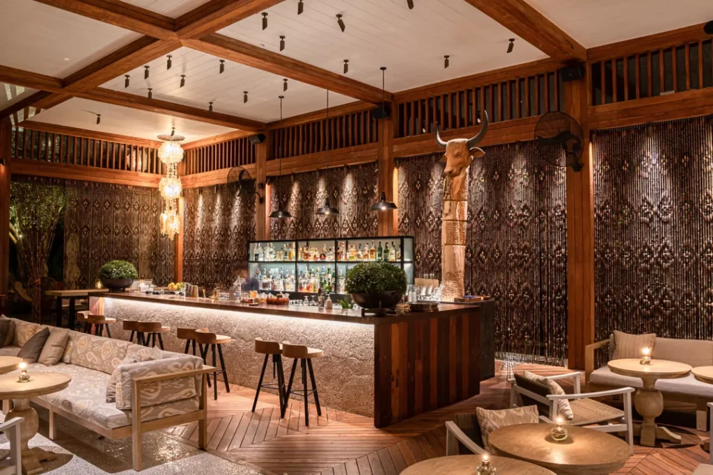 A cozy, upscale bar with a wooden interior, cushioned seating, warm lighting, and decorative beaded curtains. A well-stocked bar stands in the center, with a carved wooden bull head statue nearby.